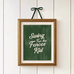 May include: A framed print with the text "Swing for the Fences Kid" in white script on a green background. The frame is gold, and a dark green ribbon is tied at the top for hanging. The print is displayed against a white, wood-panelled wall.