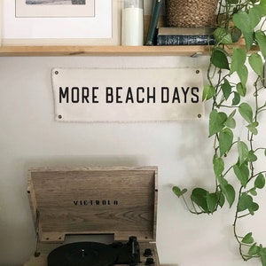K&ouml;nnte beinhalten: Ein Plattenspieler im Vintage-Stil mit dem Markennamen "VICTROLA" steht auf einer Holzoberfl&auml;che. Dar&uuml;ber h&auml;ngt ein Banner mit der Aufschrift "MORE BEACH DAYS". Ein gerahmtes Strandbild und eine gr&uuml;ne Rankpflanze erg&auml;nzen die K&uuml;stendekoration.