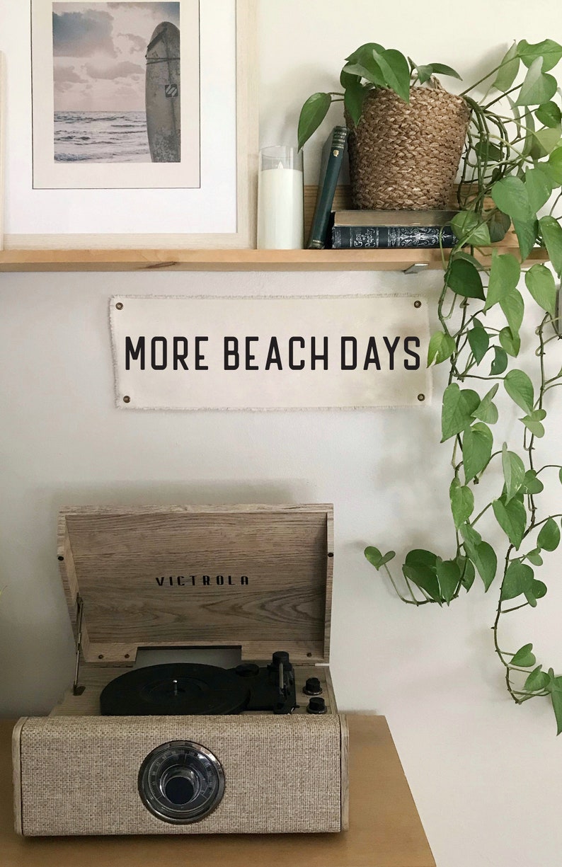 K&ouml;nnte beinhalten: Ein Plattenspieler im Vintage-Stil mit dem Markennamen "VICTROLA" steht auf einer Holzoberfl&auml;che. Dar&uuml;ber h&auml;ngt ein Banner mit der Aufschrift "MORE BEACH DAYS". Ein gerahmtes Strandbild und eine gr&uuml;ne Rankpflanze erg&auml;nzen die K&uuml;stendekoration.