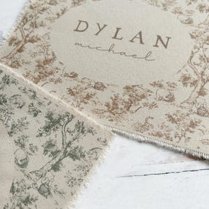 May include: A beige fabric placemat with a brown and green toile pattern. The placemat has a frayed edge and features the name "DYLAN michael" printed in brown.