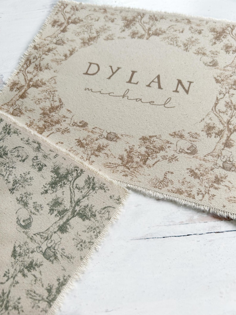 May include: A beige fabric placemat with a brown and green toile pattern. The placemat has a frayed edge and features the name "DYLAN michael" printed in brown.