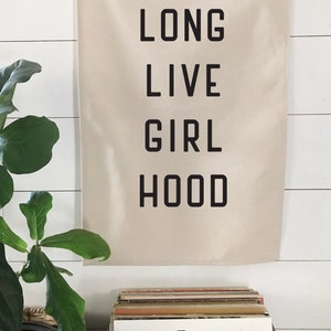 以下が含まれることがあります： 白い壁に、黒い文字で「LONG LIVE GIRL HOOD」と書かれたベージュ色のバナーが掛けられています。その下には、Fleetwood Macの「Rumours」のアルバムカバーなど、レコードが入った木箱があります。