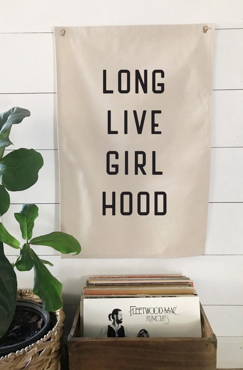 以下が含まれることがあります： 白い壁に、黒い文字で「LONG LIVE GIRL HOOD」と書かれたベージュ色のバナーが掛けられています。その下には、Fleetwood Macの「Rumours」のアルバムカバーなど、レコードが入った木箱があります。