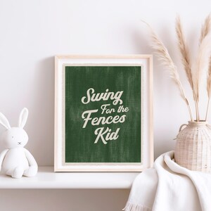 May include: A framed print with the text "Swing for the Fences Kid" in white script on a dark green background. The print is in a light wood frame and is displayed on a white shelf with a white bunny figurine and a vase of dried plants.