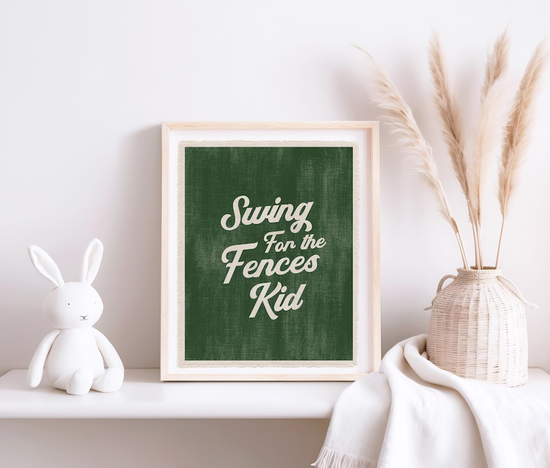 May include: A framed print with the text "Swing for the Fences Kid" in white script on a dark green background. The print is in a light wood frame and is displayed on a white shelf with a white bunny figurine and a vase of dried plants.