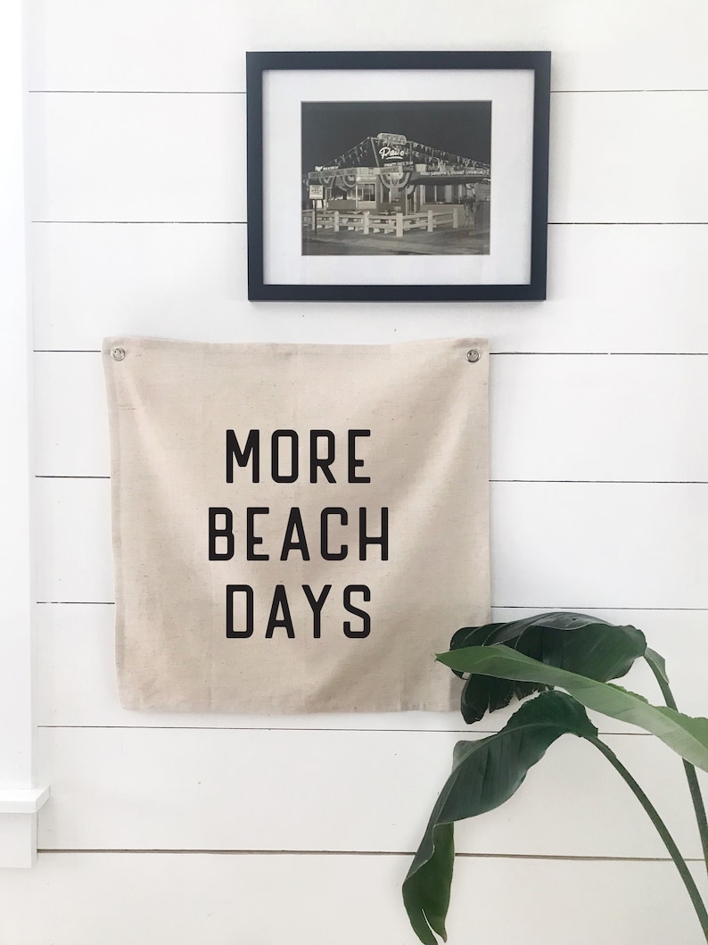 Puede incluir: Una pancarta de lona blanca con texto negro que dice "MORE BEACH DAYS" colgada en una pared blanca de listones de madera. Una foto enmarcada en negro de un edificio con luces est&aacute; encima de la pancarta.