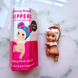 K&ouml;nnte beinhalten: Eine dekorative Miniaturfigur der Marke Sonny Angel. Die Figur ist nackt mit einem wei&szlig;en und orangefarbenen Hut. Auf der Verpackung steht "Sonny Angel HIPPERS" und "Looking Back Series".