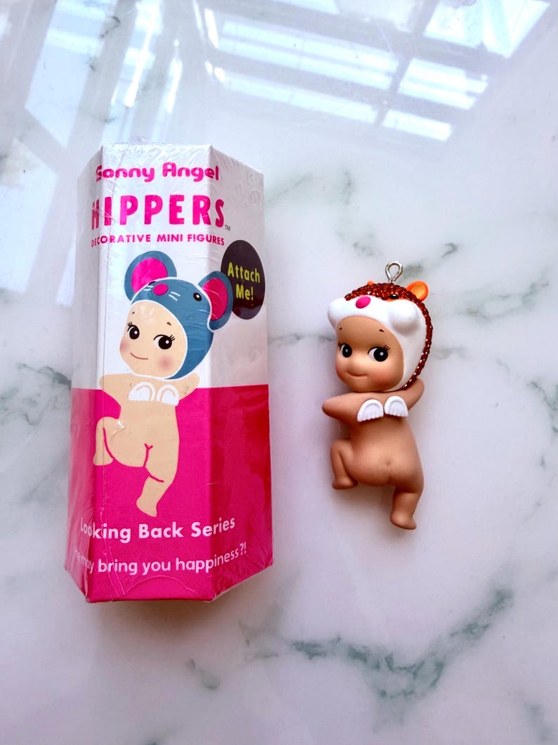 K&ouml;nnte beinhalten: Eine dekorative Miniaturfigur der Marke Sonny Angel. Die Figur ist nackt mit einem wei&szlig;en und orangefarbenen Hut. Auf der Verpackung steht "Sonny Angel HIPPERS" und "Looking Back Series".