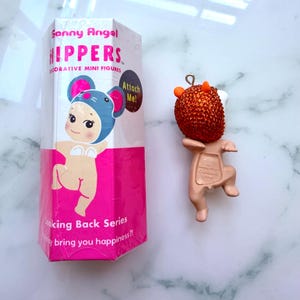 K&ouml;nnte beinhalten: Eine Sonny Angel "Hippers" dekorative Miniaturfigur, mit einer Box mit einer Cartoonfigur in einem M&auml;usekost&uuml;m. Die Figur ist eine kleine, hellbraune Figur mit einem orangefarbenen, strukturierten Kopf und kleinen H&ouml;rnern. Auf der Box steht "Looking Back Series".