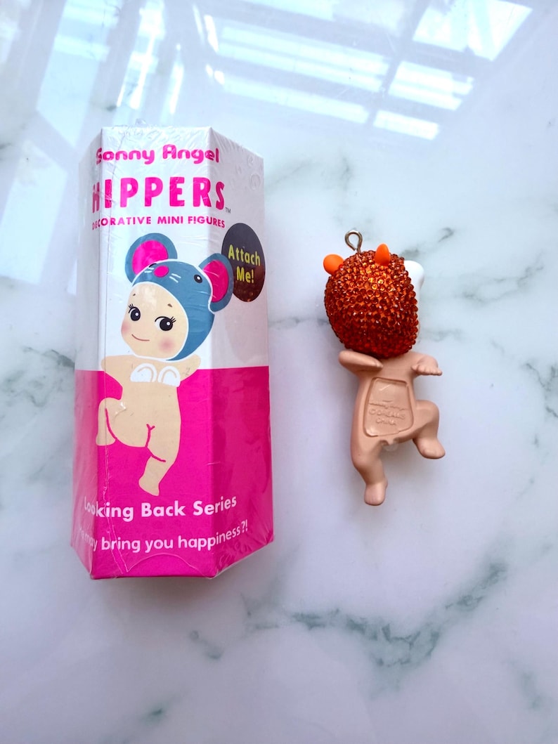 K&ouml;nnte beinhalten: Eine Sonny Angel "Hippers" dekorative Miniaturfigur, mit einer Box mit einer Cartoonfigur in einem M&auml;usekost&uuml;m. Die Figur ist eine kleine, hellbraune Figur mit einem orangefarbenen, strukturierten Kopf und kleinen H&ouml;rnern. Auf der Box steht "Looking Back Series".