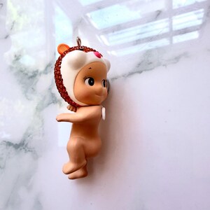 K&ouml;nnte beinhalten: Kleine, nacktfarbene Babypuppe mit einem wei&szlig;en und rotbraunen Hut im Igel-Design. Der Hut ist mit rotbraunen Strasssteinen verziert. Die Figur hat ein kleines, rosa Herz auf der Stirn und eine silberne Schlaufe zum Aufh&auml;ngen.