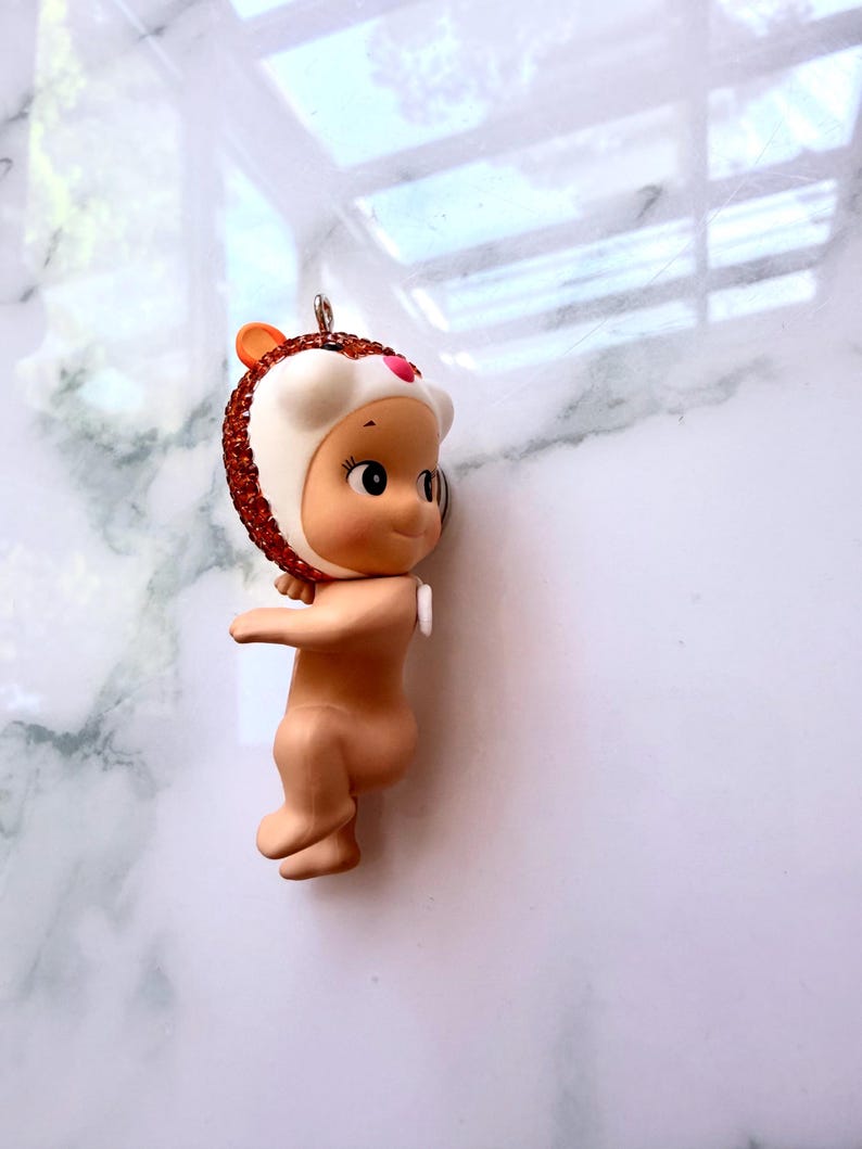 K&ouml;nnte beinhalten: Kleine, nacktfarbene Babypuppe mit einem wei&szlig;en und rotbraunen Hut im Igel-Design. Der Hut ist mit rotbraunen Strasssteinen verziert. Die Figur hat ein kleines, rosa Herz auf der Stirn und eine silberne Schlaufe zum Aufh&auml;ngen.