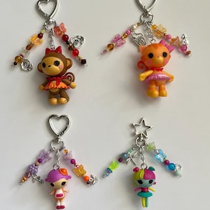 May include: Four keychains featuring small plastic character figures. Each keychain has a metal heart or star clasp, colourful beads, and charms shaped like butterflies, flowers, and insects. The figures include a brown monkey, an orange cat, and two dolls.