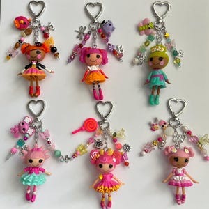 May include: Six doll keychains with heart-shaped clasps, each featuring a small plastic doll with yarn-like hair, colourful dresses, and dangling charms including beads, butterflies, flowers, and small animals in various bright colour palettes.