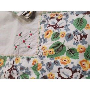 May include: A patchwork textile featuring floral patterns and embroidered details. The design includes yellow and orange roses, green leaves, and small blue flowers on a white background. The textile also has embroidered pink flowers.