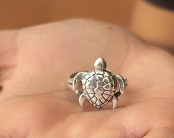 May include: A silver-colored ring in the shape of a sea turtle. The ring features a detailed turtle design with a textured shell and flippers. The ring is shown on a person's open palm.