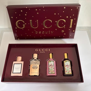 May include: A Gucci Beauty gift set featuring four miniature perfume bottles in a deep burgundy box with gold star accents. The set includes Gucci Bloom, Gucci Guilty, and two Gucci Flora fragrances, all displayed in a matching burgundy tray.
