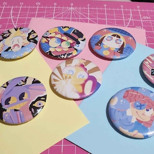 May include: A selection of colourful, round and oval buttons featuring cartoon character designs. The buttons are arranged on pink and pastel-coloured paper, creating a vibrant display. The designs feature a variety of colours and styles.