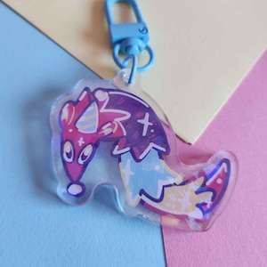 May include: A clear acrylic keyring with a cartoon creature design, wearing a party hat and a cross. The creature is mainly purple, pink, and blue, with a blue clasp. The background is a mix of blue, yellow, and pink.