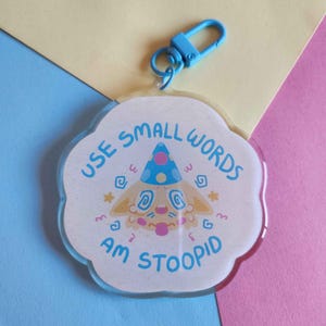 Könnte beinhalten: Ein klarer, wolkenförmiger Schlüsselanhänger mit einem Cartoon-Hund mit Partyhut. Der Schlüsselanhänger hat den Text "USE SMALL WORDS AM STOOPID" in Hellblau. Ein blauer Verschluss ist angebracht.