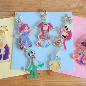 May include: A collection of nine clear acrylic charms featuring colourful cartoon character illustrations. Each charm is attached to a gold-toned star-shaped clasp. The characters include various figures in vibrant outfits, including jesters and animals.