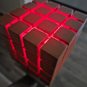 May include: A geometric cube-shaped lamp with a warm brown finish. The cube is composed of smaller square blocks, with vibrant red light emanating from the spaces between them. The lamp is a modern decorative piece.