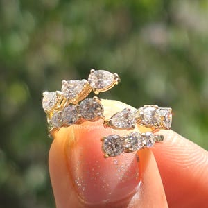 Può includere: Un anello aperto in oro con una serie di pietre trasparenti a taglio pera e rotondo disposte lungo la fascia. L'anello presenta un design aperto con il testo tabicejewelry visibile sulla superficie interna. Le pietre sono incastonate in metallo giallo.