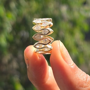 May include: A stack of gold rings, each featuring a marquise-cut diamond. The rings are held up against a blurred green background. The rings have a unique design, with the diamonds set in a repeating pattern. The rings are a bright gold color.