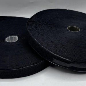 May include: Two circular spools of black hook and loop fastener tape. Each spool features a continuous strip of black fabric wound tightly around a central plastic core, displaying a textured surface characteristic of adhesive fastening materials.