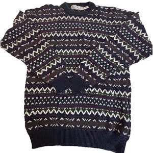 May include: A navy blue sweater with a geometric pattern in white, green, and burgundy. The sweater has a crew neck, ribbed cuffs, and a ribbed hem. The brand name is visible on the collar.