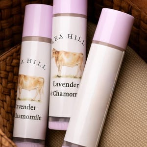 May include: Three cylindrical tubes of Lea Hill Lavender & Chamomile products. Each tube has a light purple cap and a label with a cow illustration. The tubes are arranged in a woven basket.