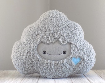 Puede incluir: Un cojín de felpa gris con forma de nube con una cara sonriente y un detalle de corazón azul. El cojín tiene un exterior suave y texturizado. Está diseñado para fines decorativos o de confort y está colocado sobre una superficie de madera blanca.