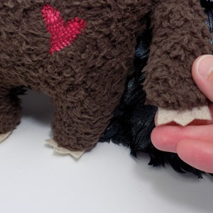 Op de afbeelding: Een bruin pluche speelgoed met een rood hartvormig lapje. Het speelgoed heeft een harige textuur en lichtgekleurde vilten voetjes. Zwart, veervormig materiaal is zichtbaar bij de voeten. Een hand houdt de voet van het speelgoed vast.
