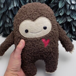 Op de afbeelding: Een bruine pluche Bigfoot-pop met een lichtbruin gezicht, zwarte knoopogen en een gestikt glimlachje. Het heeft een rood hart op de borst en kleine armen en voeten. De pop wordt in een hand gehouden.