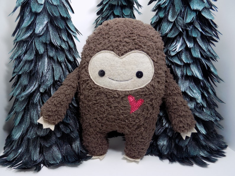 Op de afbeelding: Een bruine pluche Bigfoot-pop met een vilten gezicht, knoopogen en een gestikte glimlach. Een rood hart is op de borst geborduurd. De pop staat voor drie decoratieve, donkergroene veerbomen.