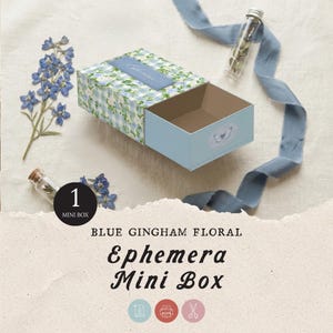 May include: A blue gingham floral Ephemera Mini Box with a light blue drawer. The box is decorated with a floral pattern and a blue ribbon. A small glass vial with dried flowers and a sprig of blue flowers are also present.