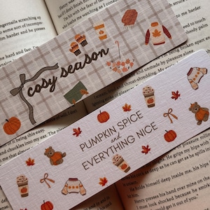 Könnte beinhalten: Zwei rechteckige Lesezeichen mit Herbstmotiven. Eines zeigt ein beiges Karomuster, den Text "cosy season" sowie Illustrationen von Schal, Pullover, Fäustlingen und Blättern. Das andere hat einen hellen Hintergrund, den Text "PUMPKIN SPICE and EVERYTHING NICE" und Symbole von Kürbissen, Bären, Pullovern und Kaffeebechern.