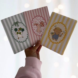 Könnte beinhalten: Drei Notizbücher mit pastellfarbenen vertikalen Streifen. Jedes Notizbuch zeigt eine andere Cartoonfigur in einem geschwungenen ovalen Rahmen. Zu den Figuren gehören eine Ente mit Kleeblättern, ein Hase mit einem Osterei und ein Bär mit Blumen.