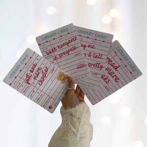 May include: A hand holds several white library cards with pink lines and text. The cards have phrases like "just one more chapter" and "book hangover in progress" written on them. A small teddy bear sticker is visible.