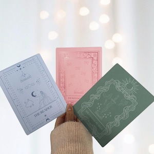 Può includere: Tre carte rettangolari con bordi decorativi e illustrazioni. Una è azzurra con un disegno di donna e luna, etichettata "THE READER". La seconda è rosa con un disegno di libro e busta, etichettata "THE ROMANCE". La terza è verde con un disegno di spada e sole, etichettata "THE FANTASY READER".