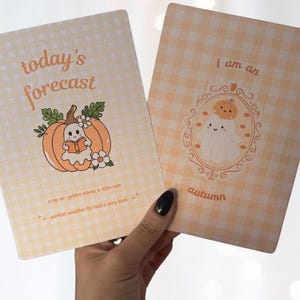 May include: Two rectangular cards with a light yellow gingham pattern. The left card features a pumpkin with a ghost reading a book and the text today's forecast, crisp air, golden leaves, a little rain, perfect weather to read a cozy book. The right card shows a ghost in an ornate frame with the text I am an autumn.