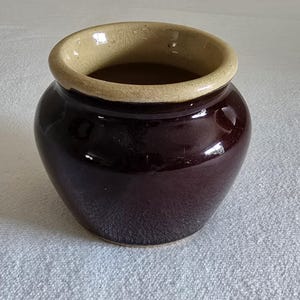 May include: A small, round, dark brown ceramic pot with a light tan rim. The pot has a glossy finish and a wide opening. This vintage-style pot is suitable for kitchen or home decor.