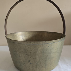 May include: A vintage brass bucket with a curved, dark metal handle. The bucket has a rounded base and a slightly flared rim. The brass has a weathered, aged appearance, suggesting it is an antique or a well-used item.