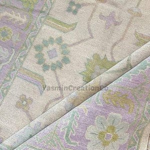Op de afbeelding: Close-up van een beige tapijt met een bloemenpatroon in tinten paars, groen en geel. Het tapijt heeft een gedetailleerd ontwerp met een gerolde rand, dat de ingewikkelde weving en textuur van de stof laat zien.