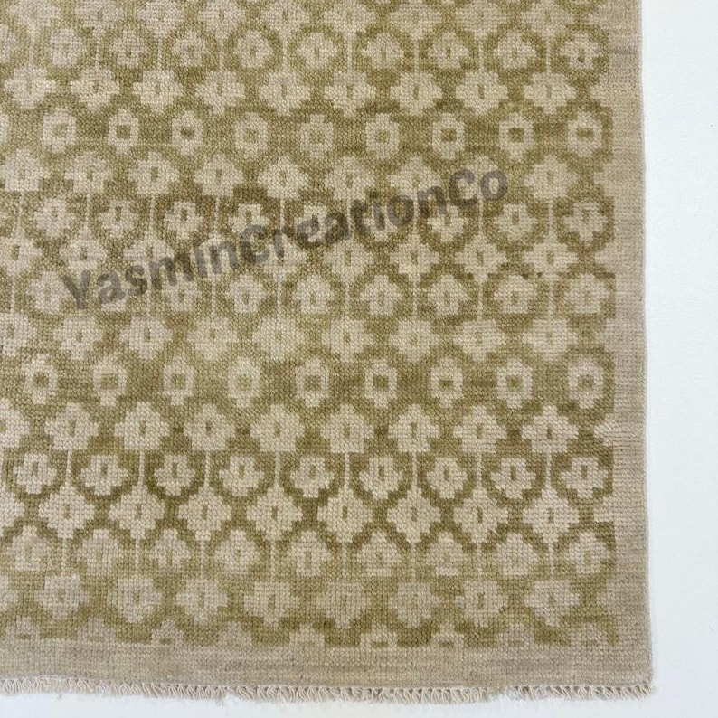 May include: A section of a rug with a repeating geometric pattern in shades of olive green and beige. The rug has a textured appearance and the text "Yasmin Creations Co" is visible.