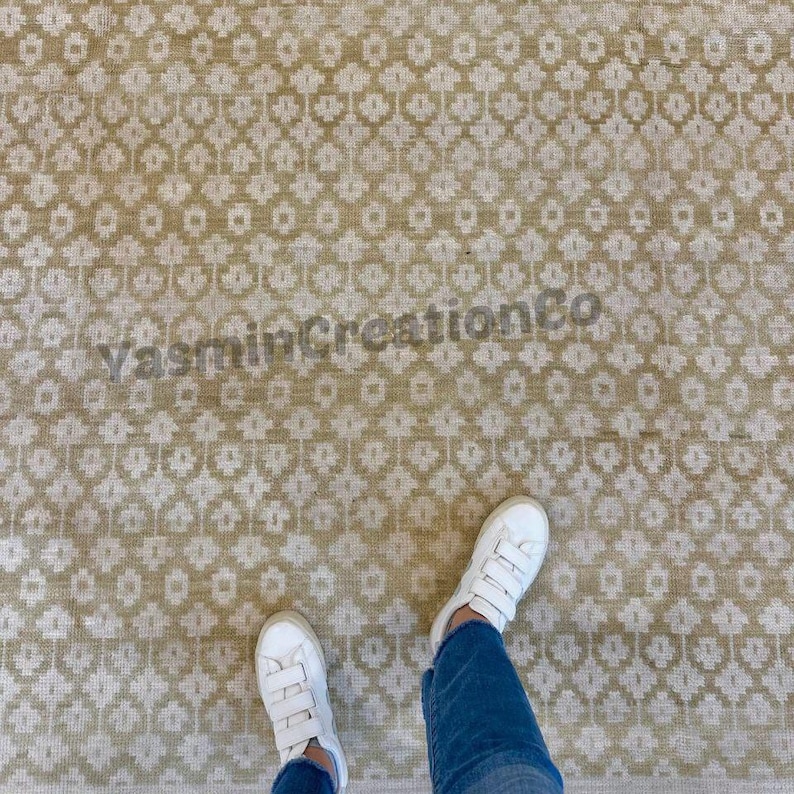 May include: Beige and off-white patterned rug with a repeating floral design. The rug has a distressed look and is seen from a top-down perspective. The text "YasminCreationCo" is visible on the rug. White sneakers and blue jeans are visible.