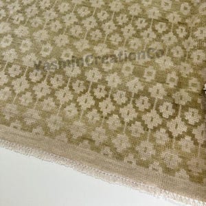 May include: Close-up of a beige and olive green patterned rug. The rug features a repeating floral design. The text "VeeMinCreationCo" is visible on the rug. The rug has a fringed edge.