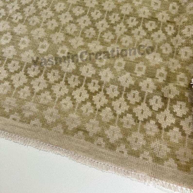 May include: Close-up of a beige and olive green patterned rug. The rug features a repeating floral design. The text "VeeMinCreationCo" is visible on the rug. The rug has a fringed edge.