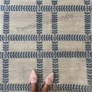 Peut inclure: Un tapis beige avec un motif de feuilles bleues en forme de grille. Le tapis a un aspect texturé. Le texte "Yasmi reationCo" est visible. Des chaussures roses sont visibles en bas de l'image.