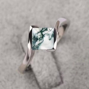 Puede incluir: Anillo de plata con una gema cuadrada que presenta un patrón único de inclusiones verdes y blancas. La banda tiene un diseño retorcido. El anillo se encuentra sobre un fondo suave y texturizado. La gema mide aproximadamente 1 cm x 1 cm.